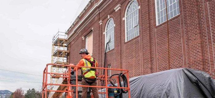A construction worker stands on a lift next to a red brick building under renovation. Scaffolding is visible against the building, and plastic covers nearby equipment.