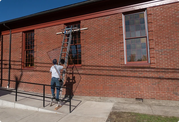 Worker positioning a WindowSLIP insert on a ladder outside a red brick building with large grid-style windows.