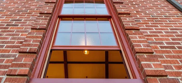 Red brick building window with glass panes and visible interior ceiling light and beams.