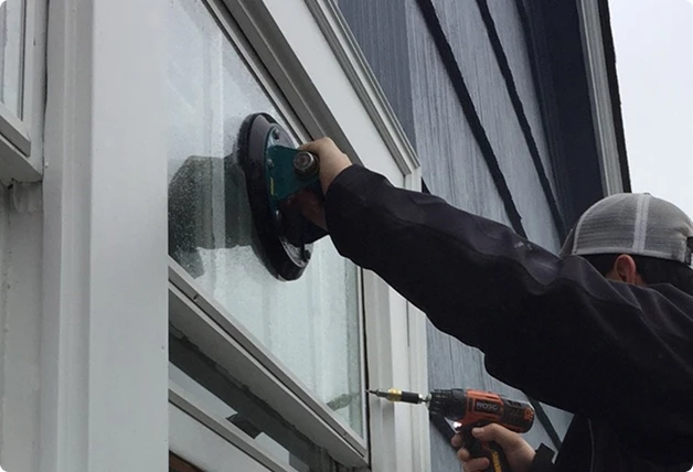 Installer securing a WindowSLIP unit on a white-framed window using a drill and suction tool against a dark gray exterior wall.